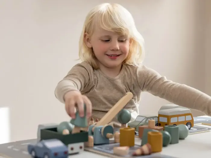 Et lille barn med lyst hår leger ved et bord med legetøjsbiler, træer og bygninger af træ, mens det smiler og fokuserer på aktiviteten.