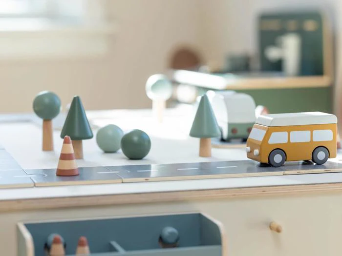 En legetøjsvej i træ med en gul bus, træer, trafikkegler og en varevogn på et legebord i et lyst, svagt oplyst rum.