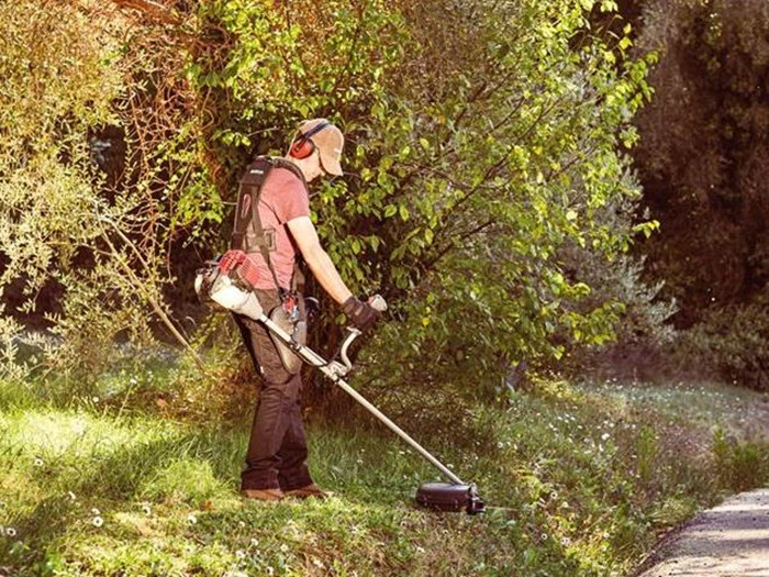 En person iført beskyttelsesudstyr bruger en græstrimmer til at slå græs på en solrig dag, omgivet af grønne buske og træer.