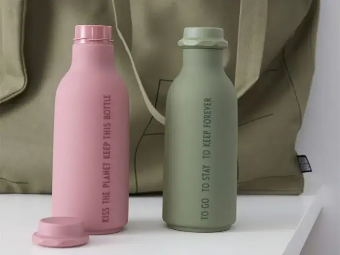 To genanvendelige vandflasker, en lyserød med teksten Kiss the planet keep this bottle og en grøn med To go to stay to keep forever, er placeret foran en beige taske.