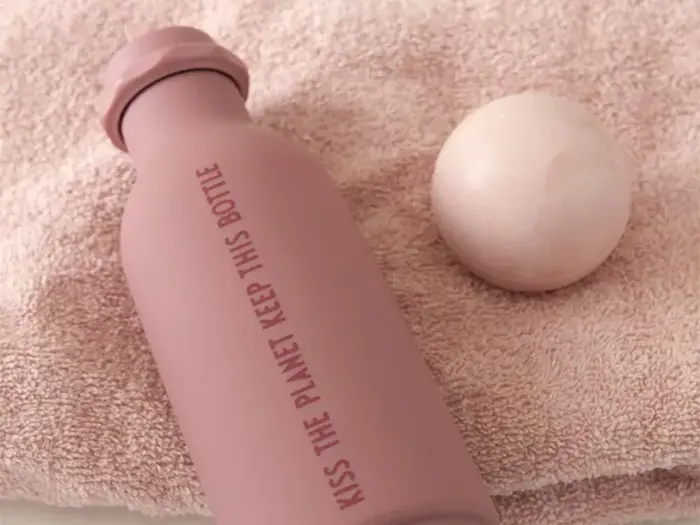 En lyserød genbrugsflaske med påskriften "Kiss the Planet Keep This Bottle" ligger på et lyserødt håndklæde ved siden af en rund badebombe.