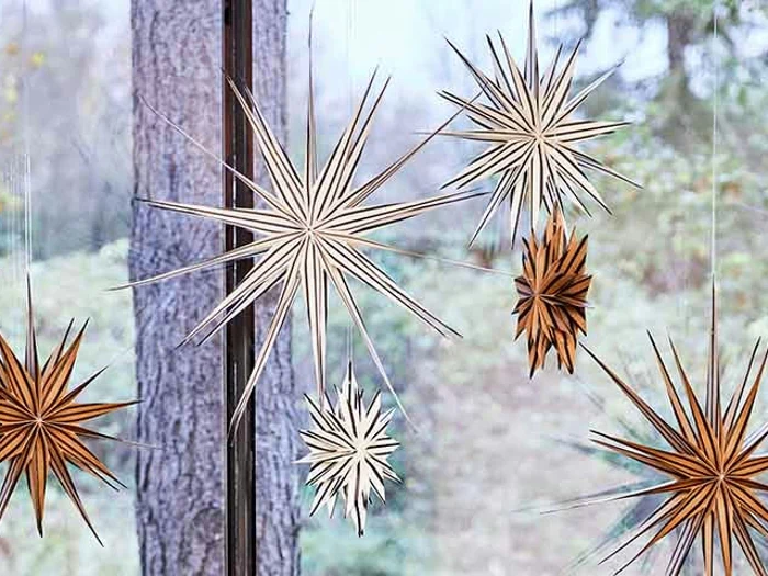 Stjerneornamenter af papir i hvidt og brunt hænger foran et vindue med en sløret udendørs skovbaggrund.