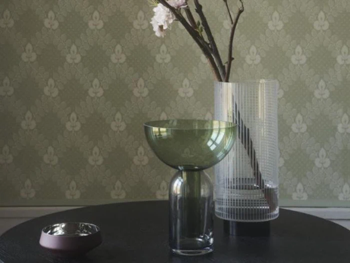 En grøn glasvase, en klar ribbet vase med blomstrende grene og to små fade står på et mørkt bord mod et mønstret grønt tapet.