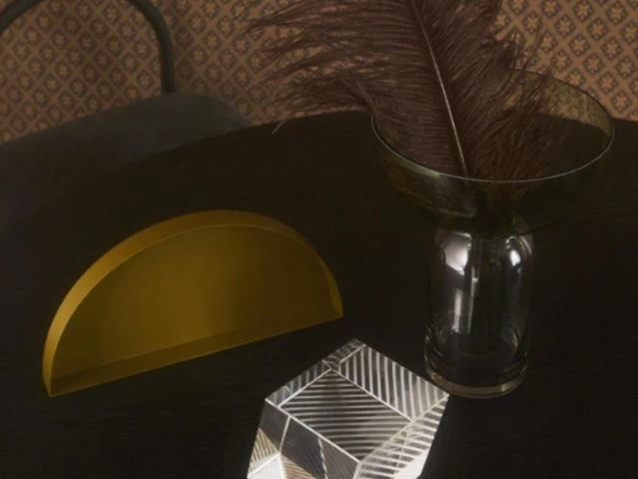 Et mørkt bord med en glasvase med en brun fjer, en gul halvcirkelformet genstand og en klar geometrisk krystal. Mønstret tapet i baggrunden.