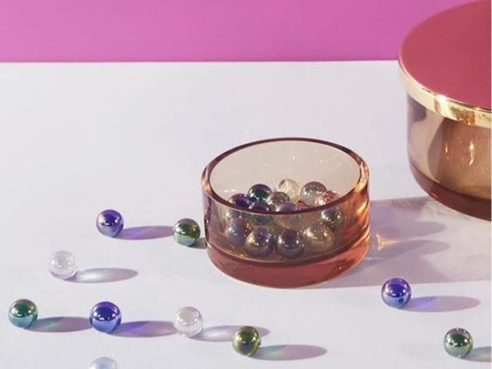 En lyserød skål fyldt med farverige glaskugler står på en hvid flade med flere kugler spredt rundt omkring og en matchende lyserød beholder ved siden af på en lyserød baggrund.