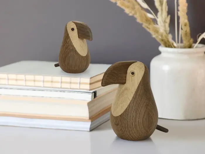 To fuglefigurer af træ står på et bord ved siden af stablede bøger og en hvid vase med tørret græs på en grå baggrund.