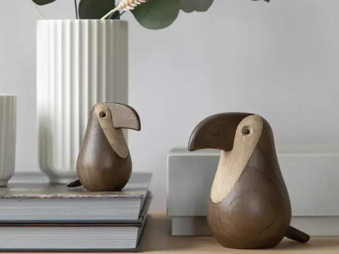 To fuglefigurer af træ med store næb står på en stak bøger og en træplade ved siden af en hvid, riflet vase med grønne blade.
