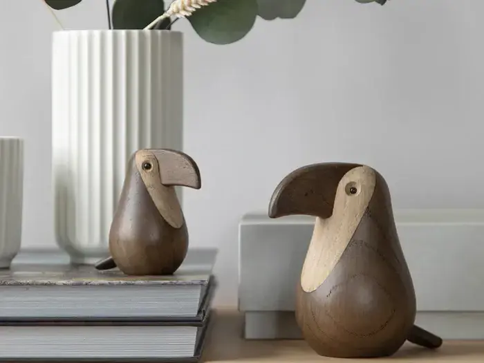 To fuglefigurer af træ med store næb sidder på stablede bøger med hvide vaser med grønne planter i baggrunden.