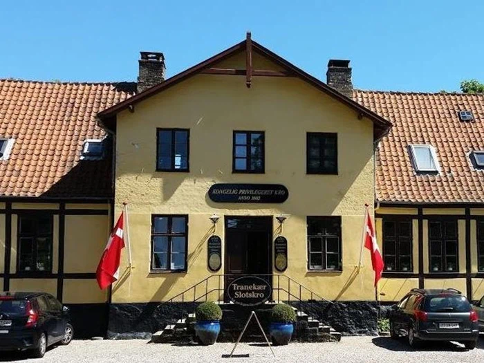 En gul bygning med rødt tegltag, to danske flag ved indgangen og et skilt med teksten Tranekær Slotskro. To biler holder parkeret foran under en klar blå himmel.