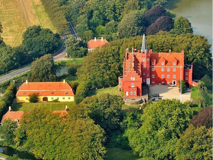 Luftfoto af et stort slot af røde mursten med tårne, omgivet af træer, en dam og nærliggende bygninger med gule tage i et frodigt grønt landskab.