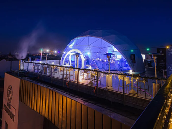 En bar på taget om aftenen har en glødende geodætisk kuppel oplyst med blå lys, udendørs siddepladser og lyskæder langs rækværket med udsigt over byens skyline.