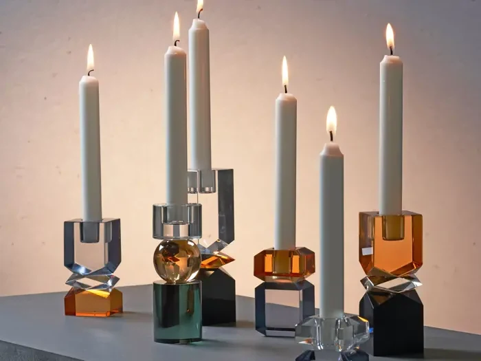 Seks hvide stearinlys i geometriske, flerfarvede glasholdere er tændt og arrangeret på en grå overflade mod en neutral baggrund.