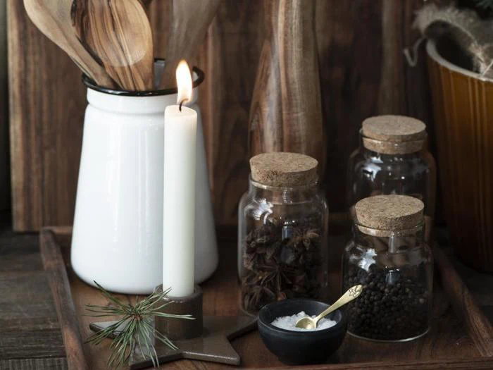 Et tændt hvidt lys, en hvid beholder med træredskaber, tre krydderikrukker med kork, en lille skål med salt og en fyrrekvist på en træbakke.