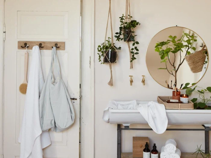 Et lyst badeværelse med håndklæder og en taske hængende på døren, potteplanter, et rundt spejl, toiletartikler på en bakke og hvide vægge, der skaber en naturlig og frisk atmosfære.