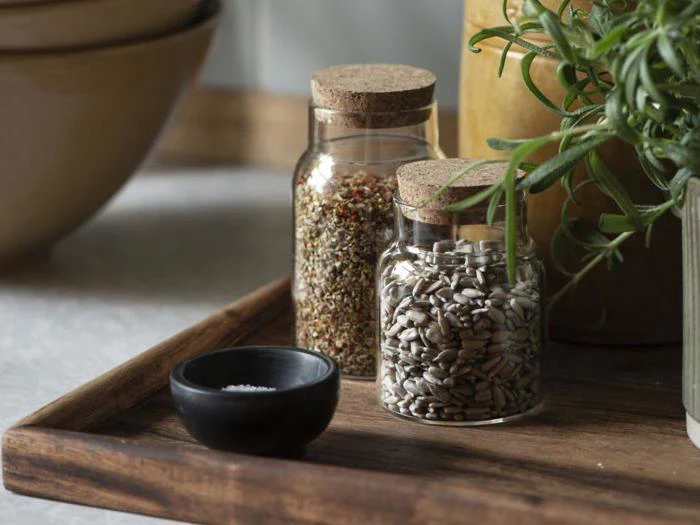 To glaskrukker med korklåg, den ene fyldt med frø og den anden med krydderier, står på en træbakke ved siden af en lille sort skål og et bundt friske, grønne krydderurter.