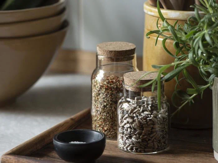 To glaskrukker med korklåg, den ene fyldt med frø og den anden med tørrede krydderurter, står på en træbakke ved siden af en sort skål og frisk rosmarin.