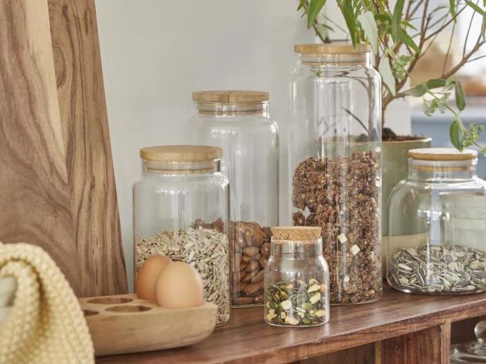 Fem glaskrukker med trælåg indeholder forskellige frø, nødder og granola. To æg og en træplade står i nærheden, alt sammen arrangeret på en træflade med en plante i baggrunden.