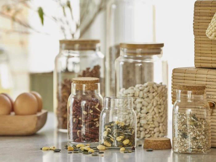 Glaskrukker fyldt med forskellige tørrede bønner og frø står på køkkenbordet ved siden af en træskål med æg og flettede kurve i baggrunden.