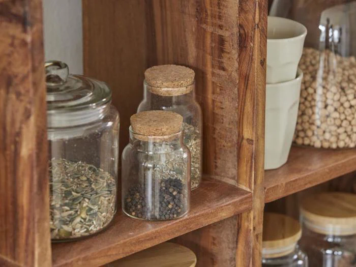 Glaskrukker med korklåg, der indeholder krydderier og frø, står pænt arrangeret på træhylder ved siden af kopper og andre krukker fyldt med tørvarer.