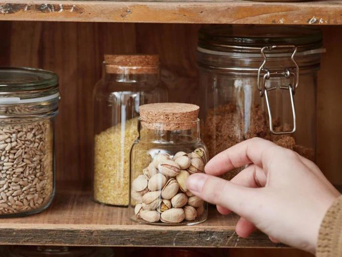 En hånd rækker ud efter en glaskrukke fyldt med pistacienødder på en træhylde ved siden af krukker med korn og frø.