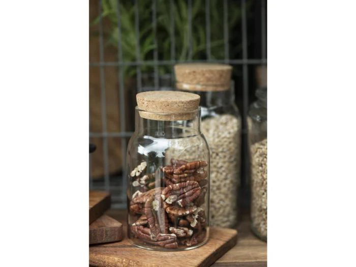 En glaskrukke med korklåg fyldt med pekannødder står på en træplade, og i baggrunden ses krukker med korn og grønne planter.