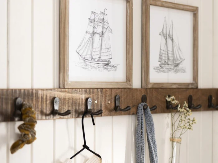 En knagerække i træ med kroge har plads til et tørklæde, en bøjle og en scrunchie. Ovenfor hænger to indrammede skibsskitser på en væg med hvide paneler; en vase med blomster står i nærheden.