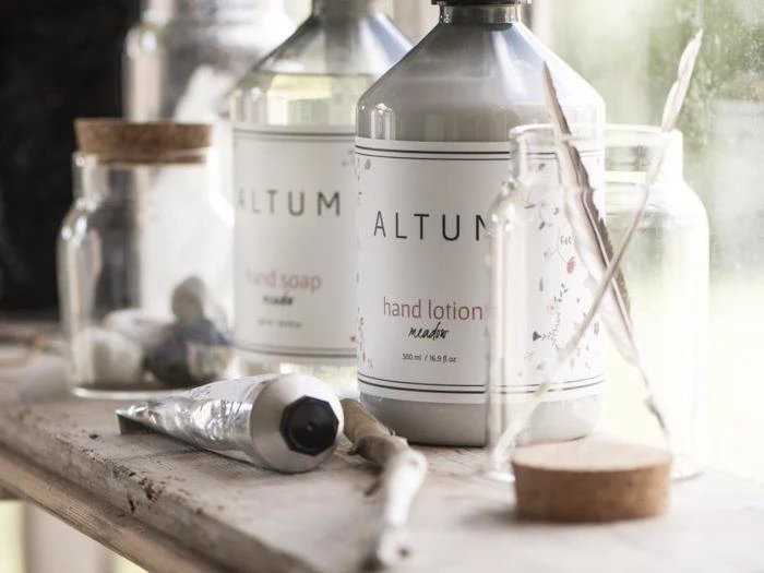 Flasker med ALTUM håndlotion og håndsæbe i en vindueskarm, omgivet af glaskrukker, en malertube og nogle pyntegenstande.