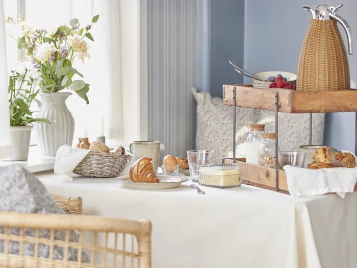 Et hyggeligt morgenbord med croissanter, brød, smør, marmelade, glas, en kaffekande og blomster i en vase i et lyst, dæmpet rum.
