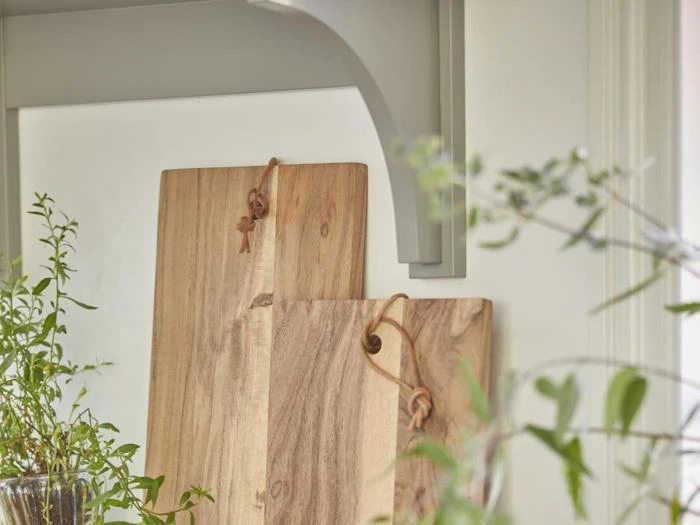 To skærebrætter af træ med læderstropper læner sig op ad en hvid væg, omgivet af grønne planter i et lyst køkken.