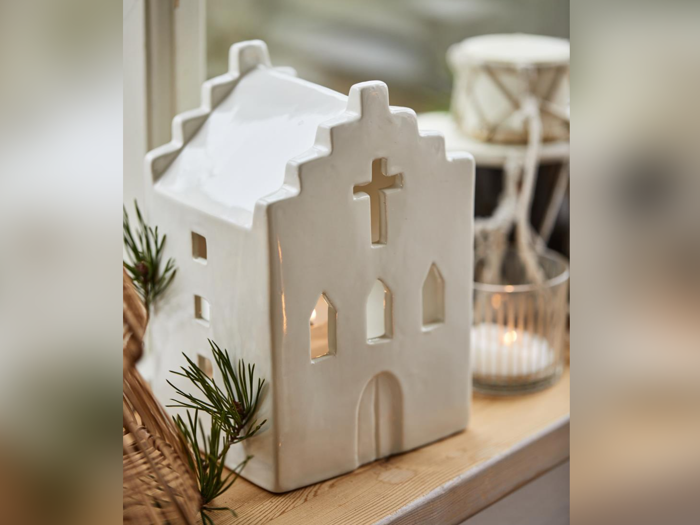 En hvid keramisk husformet lanterne med korsformede vinduer står i vindueskarmen, pyntet med grønne planter og et tændt lys ved siden af.