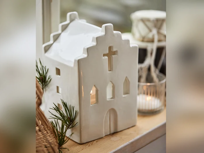 En hvid keramisk husformet lanterne med korsformede vinduer står i vindueskarmen, pyntet med grønne planter og et tændt lys ved siden af.