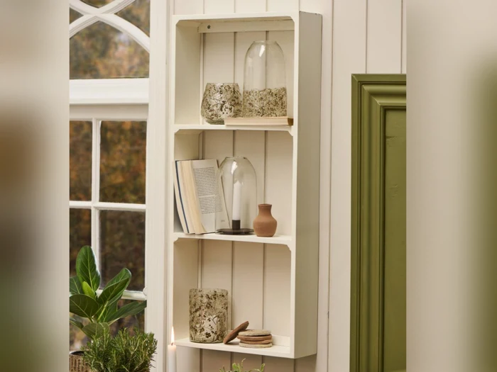 En hvid væghylde med glaskrukker, en lille lerkrukke, en stribet bog og bordskånere ved siden af et vindue og en grøn dør. En potteplante står nedenunder.