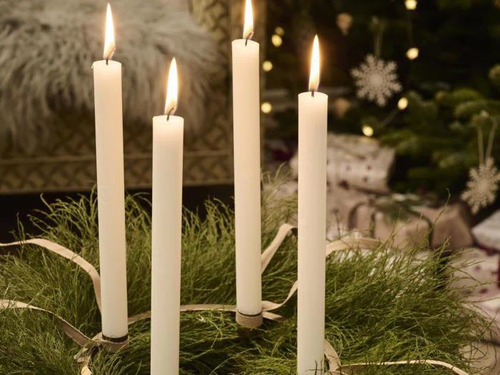 Fire tændte hvide stearinlys står i en krans af grønt løv med bånd, med et pyntet juletræ i baggrunden.