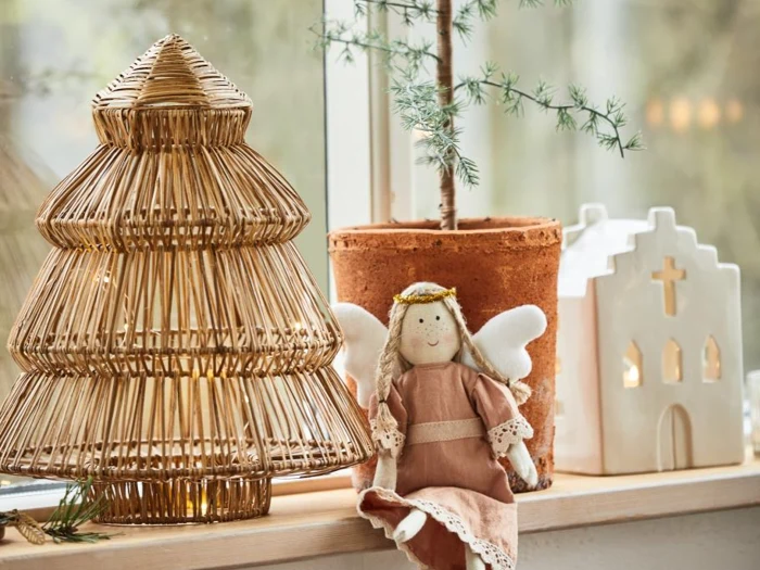 En hyggelig vindueskarm med et flettet juletræ, en potteplante, et keramikhus med et kors og en engledukke af stof i en lyserød kjole.