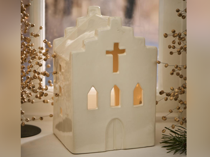 En hvid keramisk kirkeformet lysestage med oplyste vinduer står i vindueskarmen omgivet af dekorative grene og grønt.