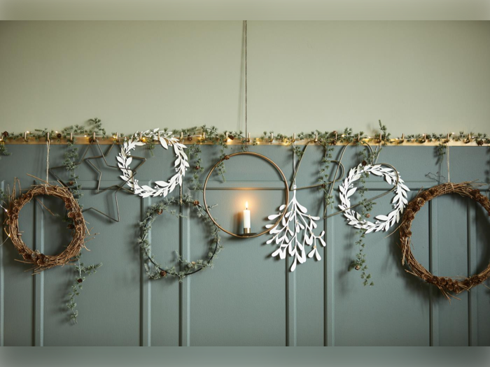 En række dekorative kranse og et lys i en guldbøjle hænger på en væg med lysegrønne paneler, hvor der er flettet grønt og kviste ind.