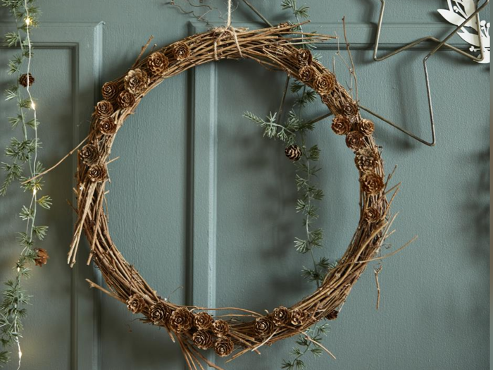 En rustik krans af kviste og små kogler hænger på en væg med blågrønne paneler, og der er grønne planter og lyskæder i nærheden.