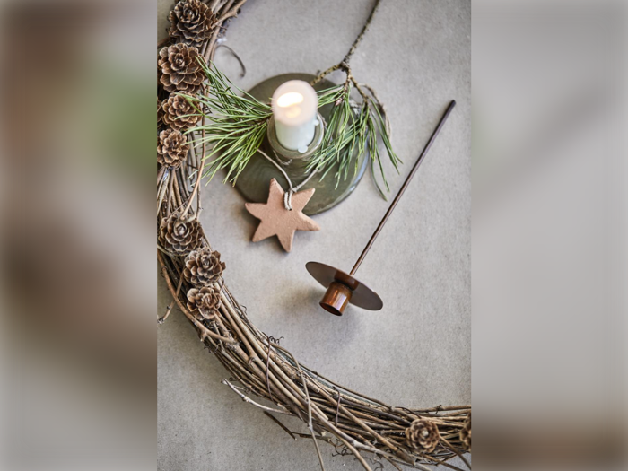 En rustik krans af kviste og små kogler, et tændt hvidt lys med fyrrekviste, et brunt stjerneornament og en brun lysestage på en lys flade.