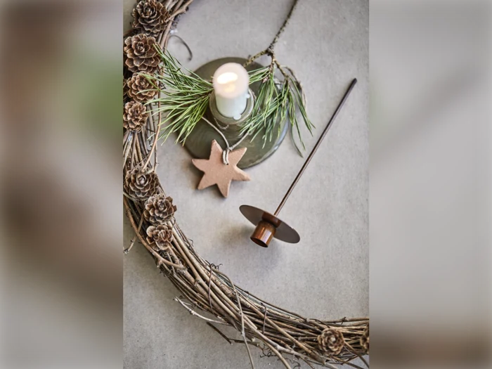 En rustik krans af kviste og små kogler, et tændt hvidt lys med fyrrekviste, et brunt stjerneornament og en brun lysestage på en lys flade.