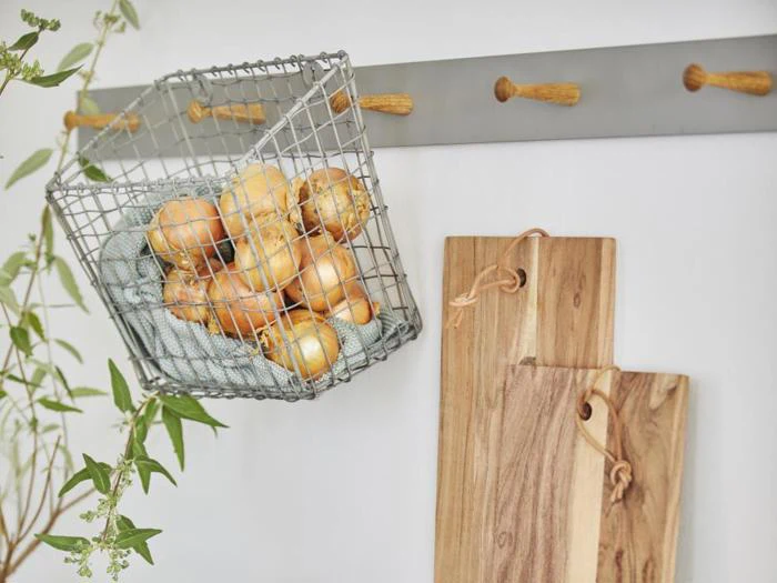 En trådkurv fyldt med løg hænger på et vægstativ ved siden af to skærebrætter af træ; en bladgren er synlig til venstre.