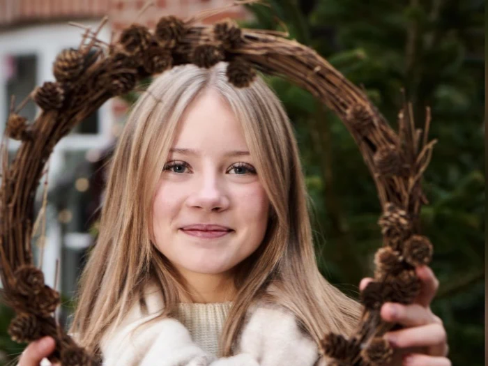 En smilende pige med blondt hår holder en cirkulær koglekrans foran sit ansigt, udendørs med grønne planter i baggrunden.