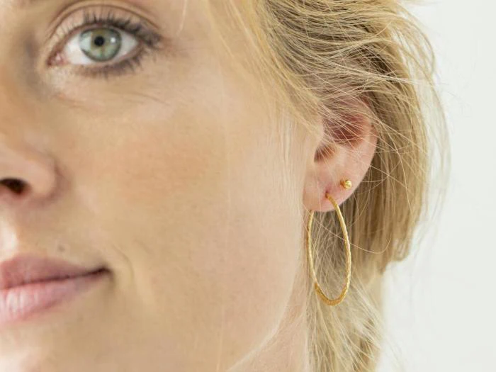 Nærbillede af en kvindes ansigt med fokus på hendes øre, der er prydet med en lille guldørering og en stor struktureret guldørering.