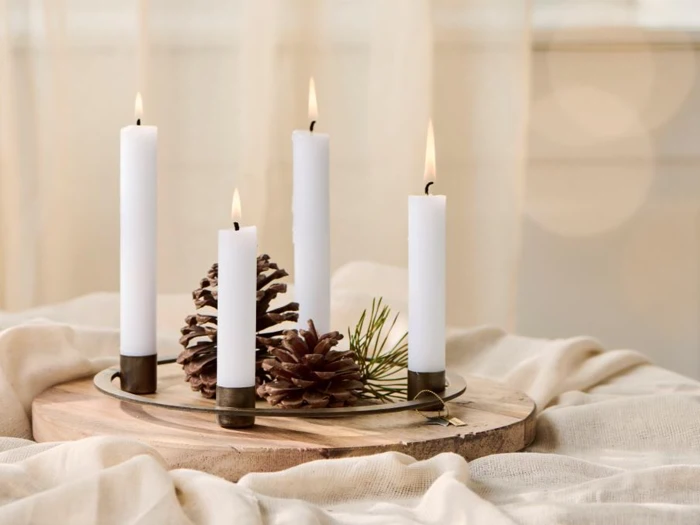 Fire tændte hvide stearinlys står oprejst på en træbakke med kogler og fyrrenåle oven på et draperet beige klæde.