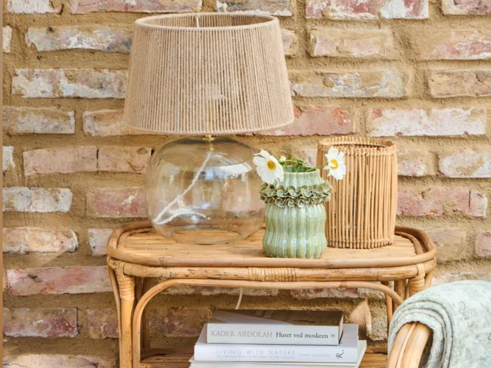 På et sidebord af rattan står en glaslampe med en skærm af jute, en grøn vase med hvide blomster, en bambuslygte og stablede bøger op ad en rustik murstensvæg.
