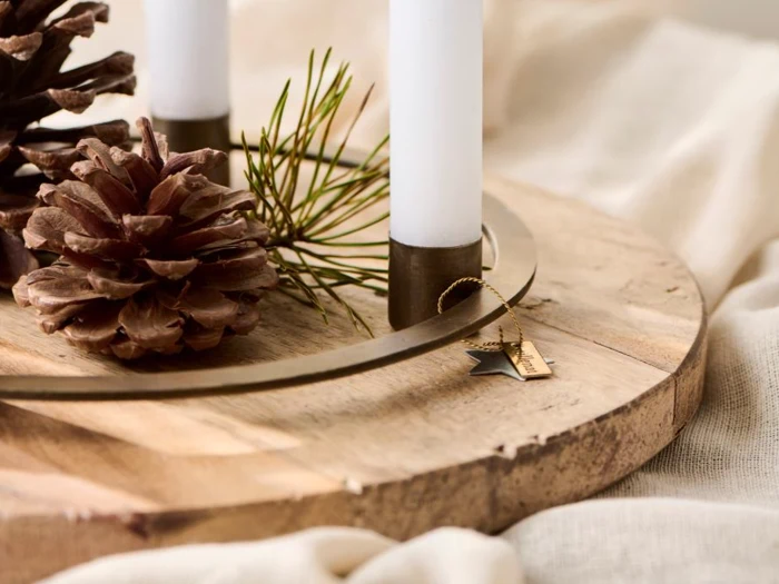 Nærbillede af en træbakke med hvide stearinlys, kogler, fyrrenåle og en lille guldstjerne på en beige stofbaggrund.