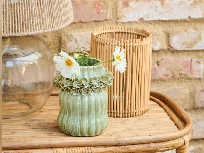En grøn keramikvase med hvide blomster står på en bambusbakke ved siden af en glaslampe med stofskærm og en bambuslygte på en baggrund af en murstensvæg.