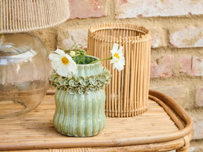 En lille grøn keramikvase med flæsedetaljer indeholder hvide blomster på et kurvebord ved siden af en glaslampe og en bambuslygte med en murstensvæg i baggrunden.