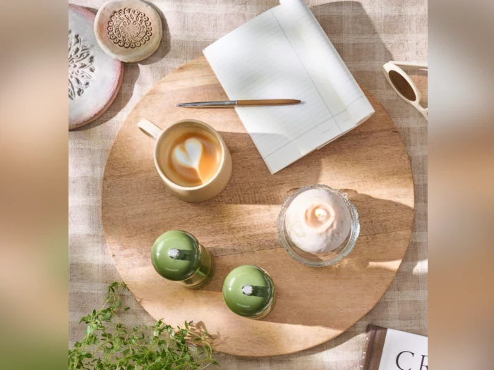 En kop latte, et wienerbrød, to grønne salt- og peberbøsser, en åben notesbog med kuglepen og solbriller på en rund træbakke, alt sammen på en ternet dug.
