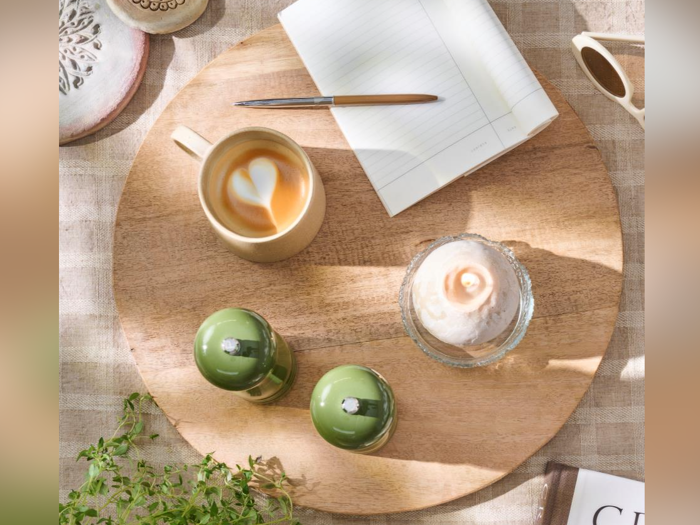 Et fladt billede af en træbakke med en latte, en notesbog med en kuglepen, en dessert, to grønne kager og lidt grønt på en beige dug.