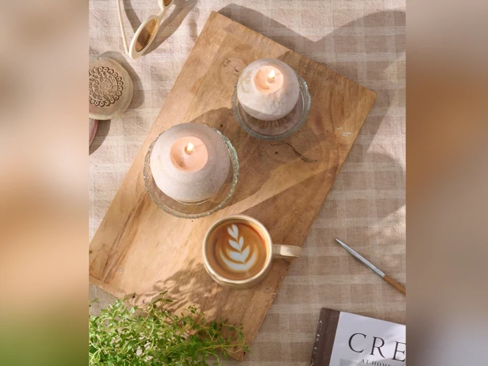 På en træplade står to tændte stearinlys og en kop latte med bladkunst ved siden af en grøn plante og en delvist synlig bog på en ternet dug.